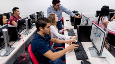 Students working in class on computers with statistical software, teacher helping one student.